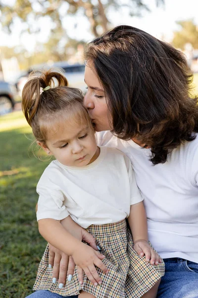 Retrato de madre morena besando mejillas de niño en camiseta blanca - foto de stock