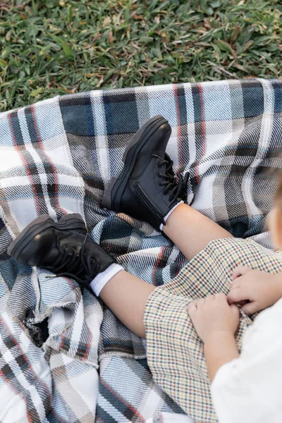 Top view of baby girl in checkered skirt and boots sitting on blanket during picnic — Stock Photo