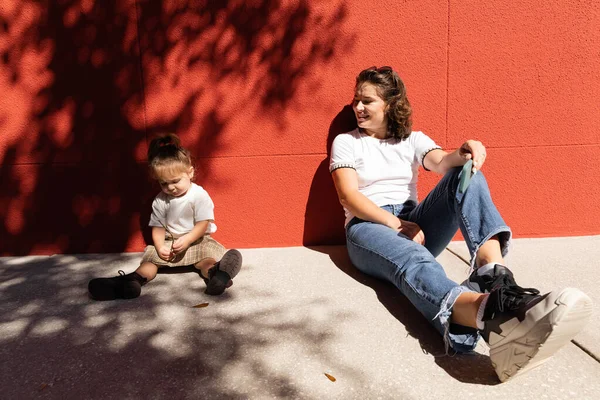 Alegre madre en jeans mirando a su hija pequeño mientras está sentado cerca de la pared roja - foto de stock
