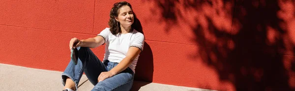 Cheerful woman in white t-shirt and blue jeans sitting near building with red wall, banner — Stock Photo