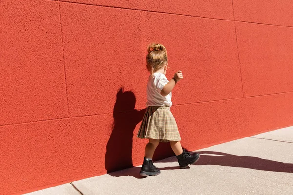 Дівчинка-малюк у картатій спідниці та білій футболці, що йде біля будівлі з червоною стіною — Stock Photo