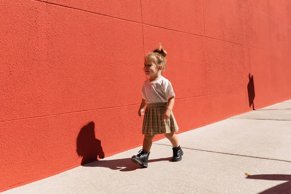 Веселая девочка в клетчатой юбке и белой футболке, идущей рядом со зданием с красной стеной — стоковое фото