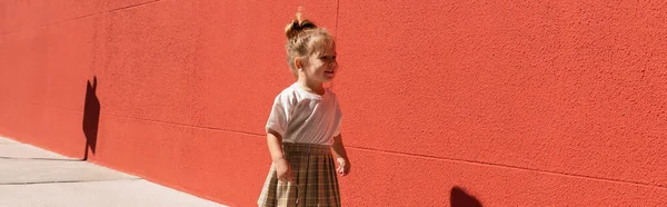 Happy toddler girl in checkered skirt and white t-shirt walking near building with red wall, banner — Stock Photo