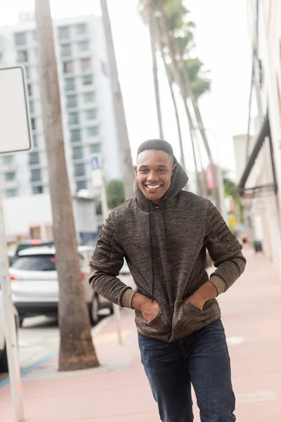 Alegre afroamericano hombre en sudadera con capucha caminando con las manos en los bolsillos en Miami - foto de stock