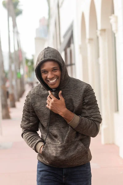 Alegre afroamericano hombre en sudadera con capucha caminando con la mano en el bolsillo en Miami - foto de stock
