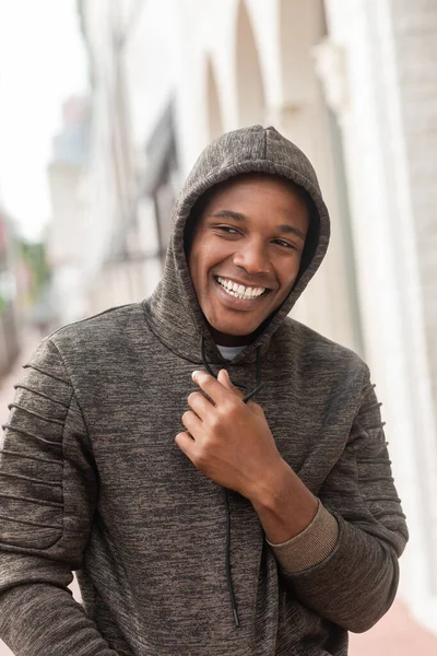 Homem americano africano alegre em camisola com capuz olhando para longe enquanto sorri ao ar livre — Fotografia de Stock