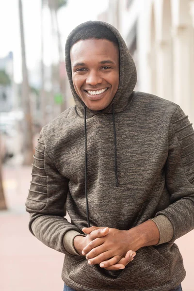 Очень радостный африканский американец в толстовке в капюшоне, смотрящий в камеру и улыбающийся на улице — стоковое фото