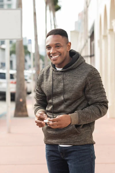 Cheerful african american man in hoodie holding earphone case on street in Miami — Stock Photo