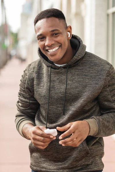 Веселий афроамериканський чоловік у светрі та бездротових навушниках, що слухають музику та тримають футляр на вулиці в Маямі — стокове фото