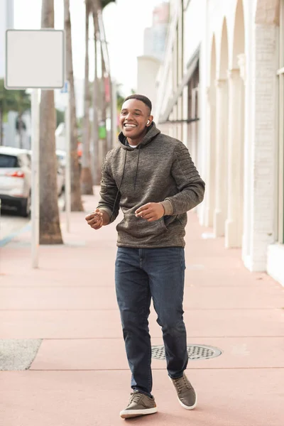 Homme afro-américain joyeux dans un écouteur sans fil écouter de la musique et marcher dans la rue à Miami — Photo de stock