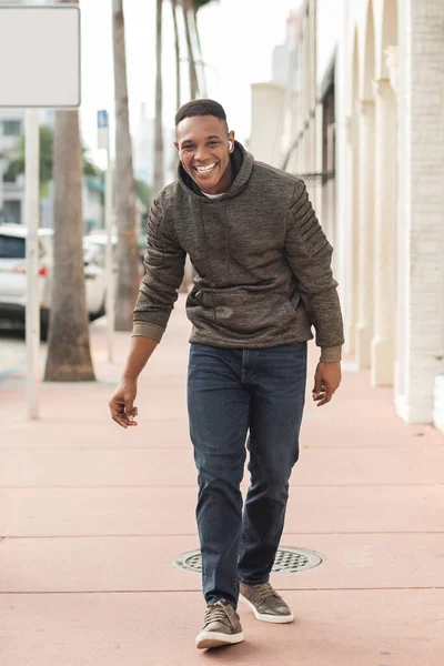 Hombre afroamericano lleno de alegría en auriculares inalámbricos caminando por la calle en Miami - foto de stock