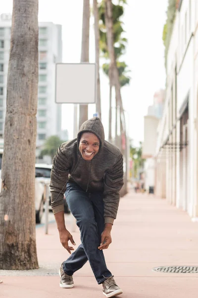 Longitud completa de hombre americano africano alegre en sudadera con capucha sonriendo al aire libre - foto de stock