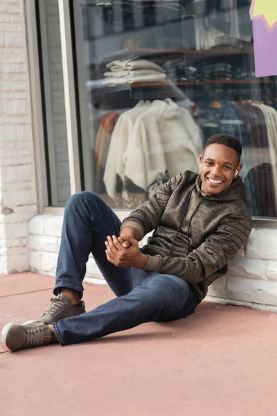 Longitud completa de alegre afroamericano hombre en sudadera con capucha sentado cerca de escaparate de tienda en Miami - foto de stock