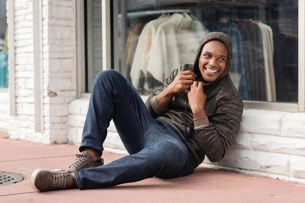Pleine longueur de joyeux homme afro-américain en sweat à capuche assis près de vitrine de magasin à Miami — Photo de stock