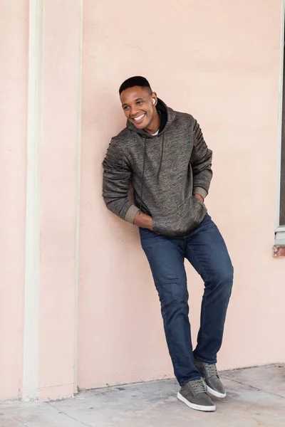 Homme afro-américain joyeux dans un écouteur sans fil debout avec les mains dans les poches près du mur — Photo de stock