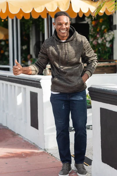 Усміхнений афроамериканський чоловік у бездротових навушниках, що висікають біля відкритого бістро — стокове фото