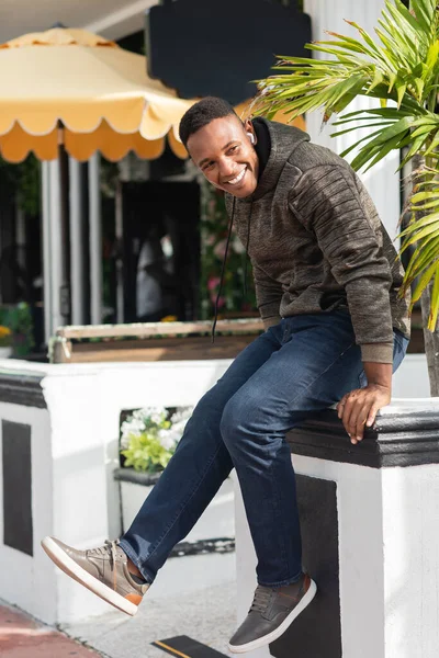 Hombre americano africano alegre en auricular inalámbrico sentado cerca de bistro al aire libre - foto de stock