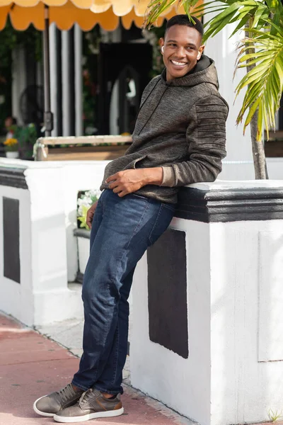 Homme afro-américain souriant dans des écouteurs sans fil debout près de bistrot extérieur — Photo de stock