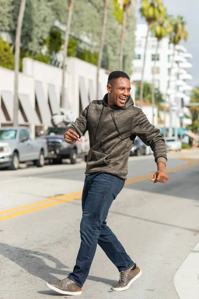 Tutta la lunghezza dell'uomo afroamericano felicissimo con cappuccio e jeans che cammina per strada a Miami — Foto stock
