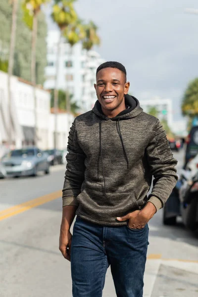 Heureux homme afro-américain en sweat à capuche et jeans marchant la main dans la poche dans la rue à Miami — Photo de stock