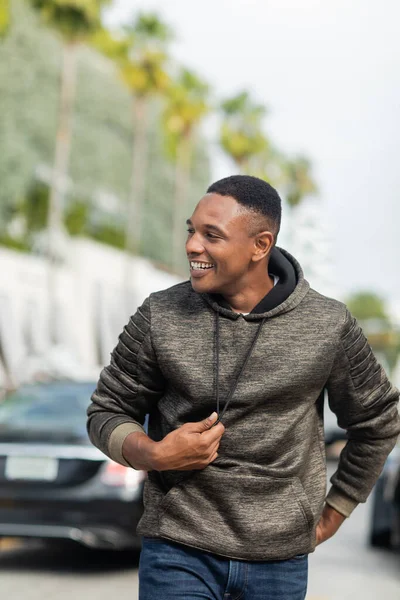 Hombre afroamericano feliz en sudadera con capucha y jeans caminando por la calle en Miami - foto de stock