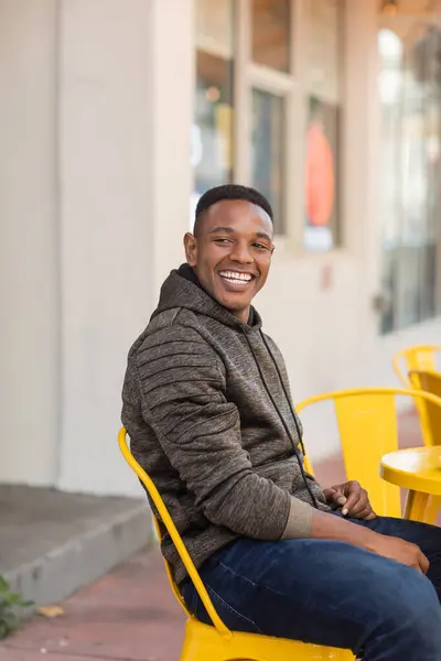 Homem americano africano alegre em capuz e jeans sentado na mesa bistro no café ao ar livre — Fotografia de Stock