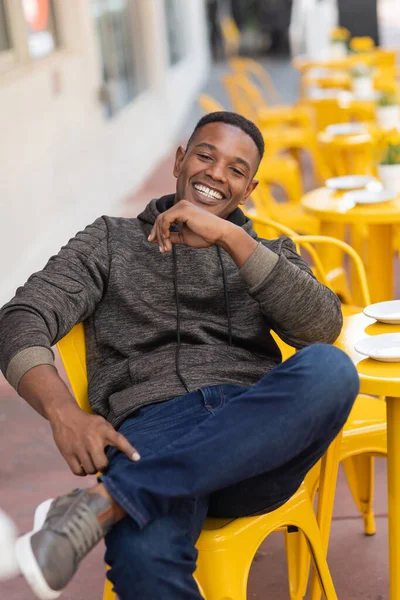 Homme afro-américain positif en sweat à capuche et jeans assis à la table de bistrot jaune dans un café extérieur — Photo de stock
