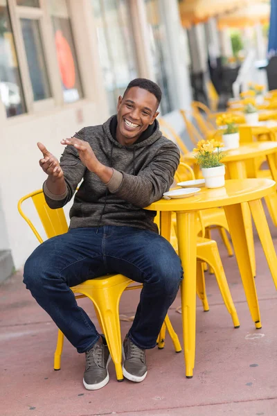 Despreocupado hombre afroamericano en sudadera con capucha y jeans sentado en la mesa del bistro en la cafetería al aire libre - foto de stock