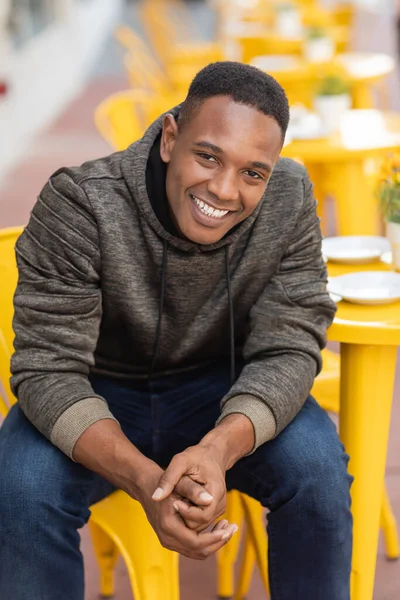 Радісний афроамериканський чоловік у светрі та джинсах, що сидить за бістро-столом у відкритому кафе — стокове фото