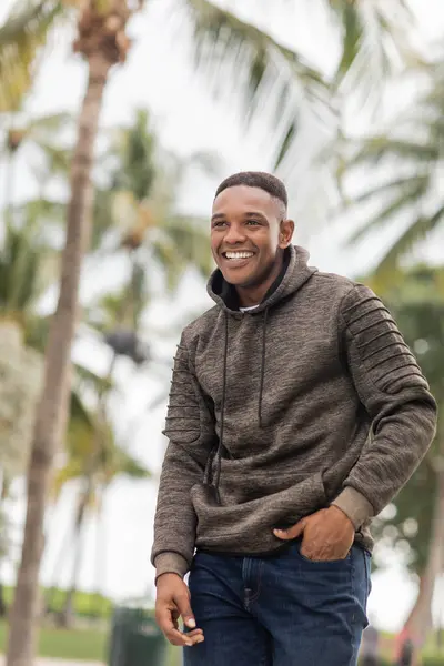 Happy african american man in hoodie standing with hand in pocket of jeans — Stock Photo