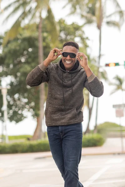Heureux homme afro-américain en sweat à capuche et jeans portant des lunettes de soleil dans la rue à Miami — Photo de stock