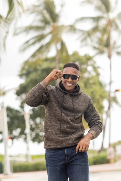 Fröhlicher afrikanisch-amerikanischer Mann in Kapuzenpulli und Jeans mit Sonnenbrille auf der Straße in Miami — Stockfoto