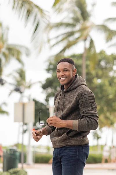 Happy african american man in hoodie and jeans holding sunglasses on street in Miami — Stock Photo