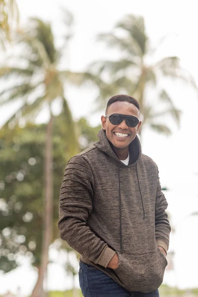 Fröhlicher afrikanisch-amerikanischer Mann in Kapuzenpulli und Sonnenbrille, der mit den Händen in den Taschen auf der Straße in Miami steht — Stockfoto