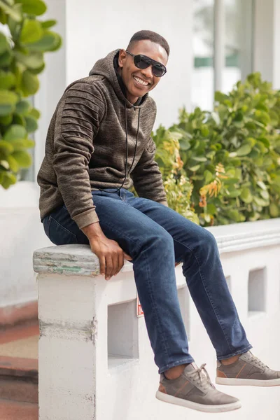 Longitud completa de hombre americano africano alegre en sudadera con capucha y gafas de sol sentado en la calle en Miami - foto de stock