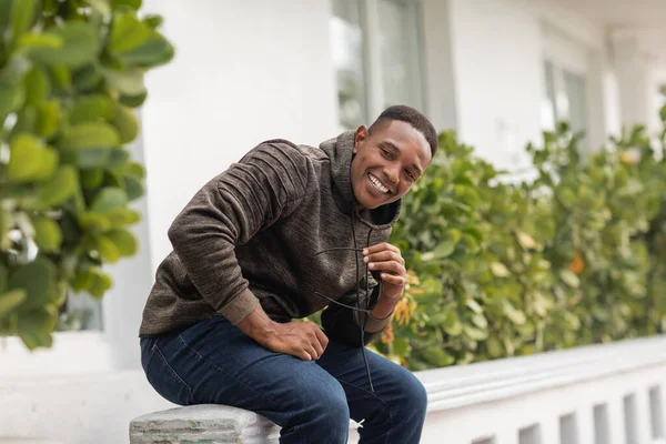 Happy african american man in hoodie holding sunglasses while sitting on street in Miami — Stock Photo