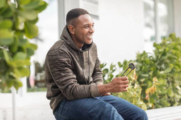 Щасливий афроамериканський чоловік в светрі і джинсах тримає сонцезахисні окуляри, сидячи на відкритому повітрі — стокове фото