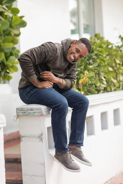 Happy african american man in hoodie and jeans holding sunglasses in teeth while sitting outdoors — Stock Photo