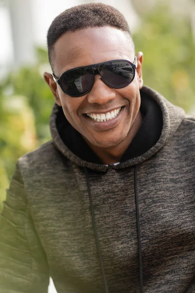Porträt eines positiven afrikanisch-amerikanischen Mannes in Kapuzenpulli und stylischer Sonnenbrille, der draußen lächelt — Stockfoto