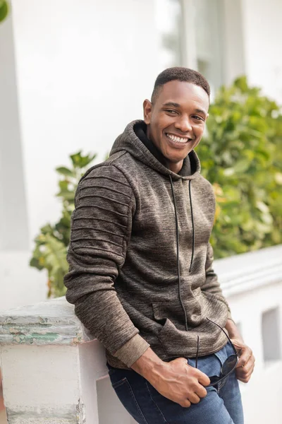 Smiling african american man in hoodie holding stylish sunglasses while standing outdoors — Stock Photo