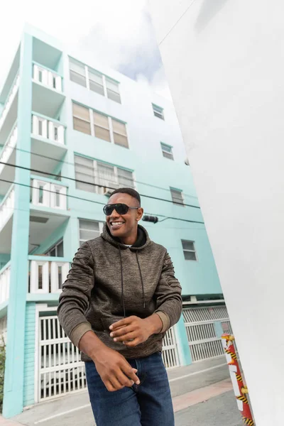 Blick auf einen glücklichen afrikanisch-amerikanischen Mann mit stylischer Sonnenbrille, der in der Nähe eines blauen Gebäudes in Miami steht — Stockfoto