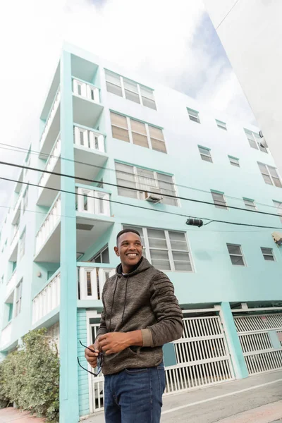 Низький кут зору щасливий афроамериканський чоловік тримає сонцезахисні окуляри, стоячи біля синьої будівлі в Маямі — стокове фото