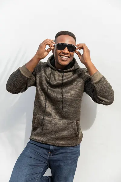 Homem americano africano feliz em camisola com capuz usando óculos de sol perto da parede branca — Fotografia de Stock