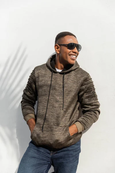 Happy african american man in sunglasses standing with hands in pockets near white wall — Stock Photo
