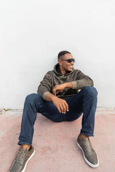 Щасливий афроамериканський чоловік у стильних сонцезахисних окулярах, що сидять біля білої стіни — стокове фото