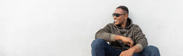 Happy african american man in stylish sunglasses sitting near white wall, banner — Stock Photo
