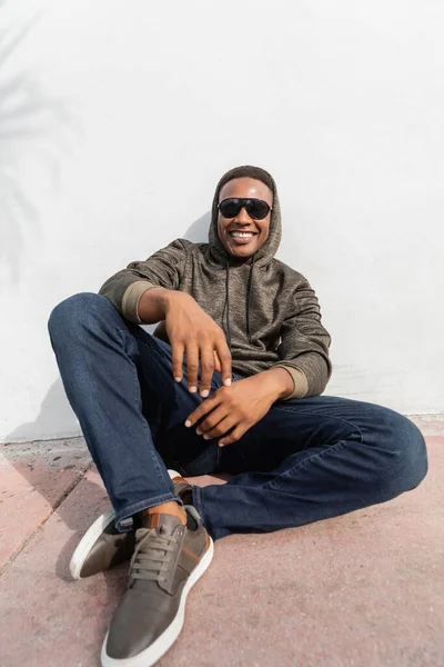Веселий афроамериканський чоловік у стильних сонцезахисних окулярах, що сидять біля білої стіни — стокове фото