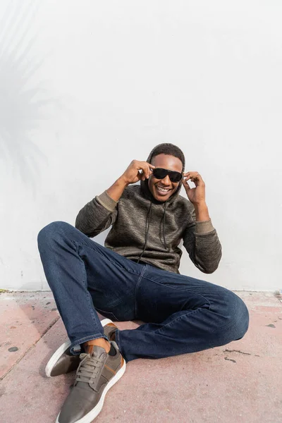 Sonriente afroamericano hombre en gafas de sol con estilo sentado cerca de la pared blanca - foto de stock