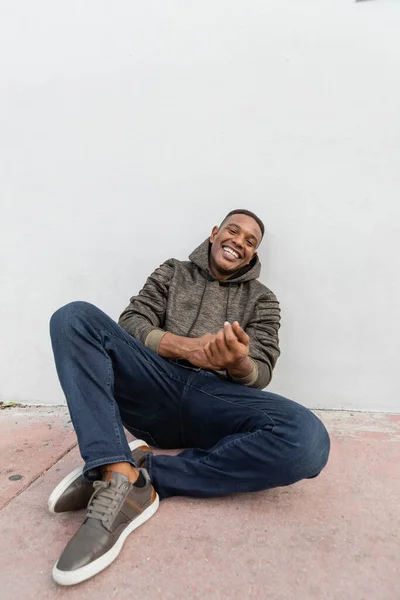 Happy african american man in jeans and hoodie smiling while sitting near white wall — Stock Photo
