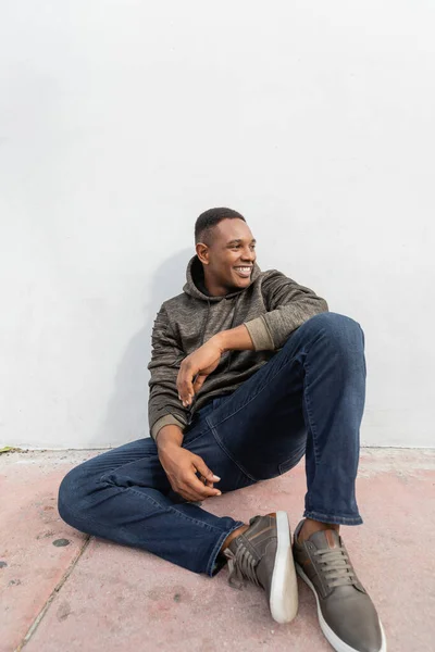Alegre afroamericano hombre en jeans y sudadera con capucha sonriendo mientras está sentado cerca de la pared blanca - foto de stock